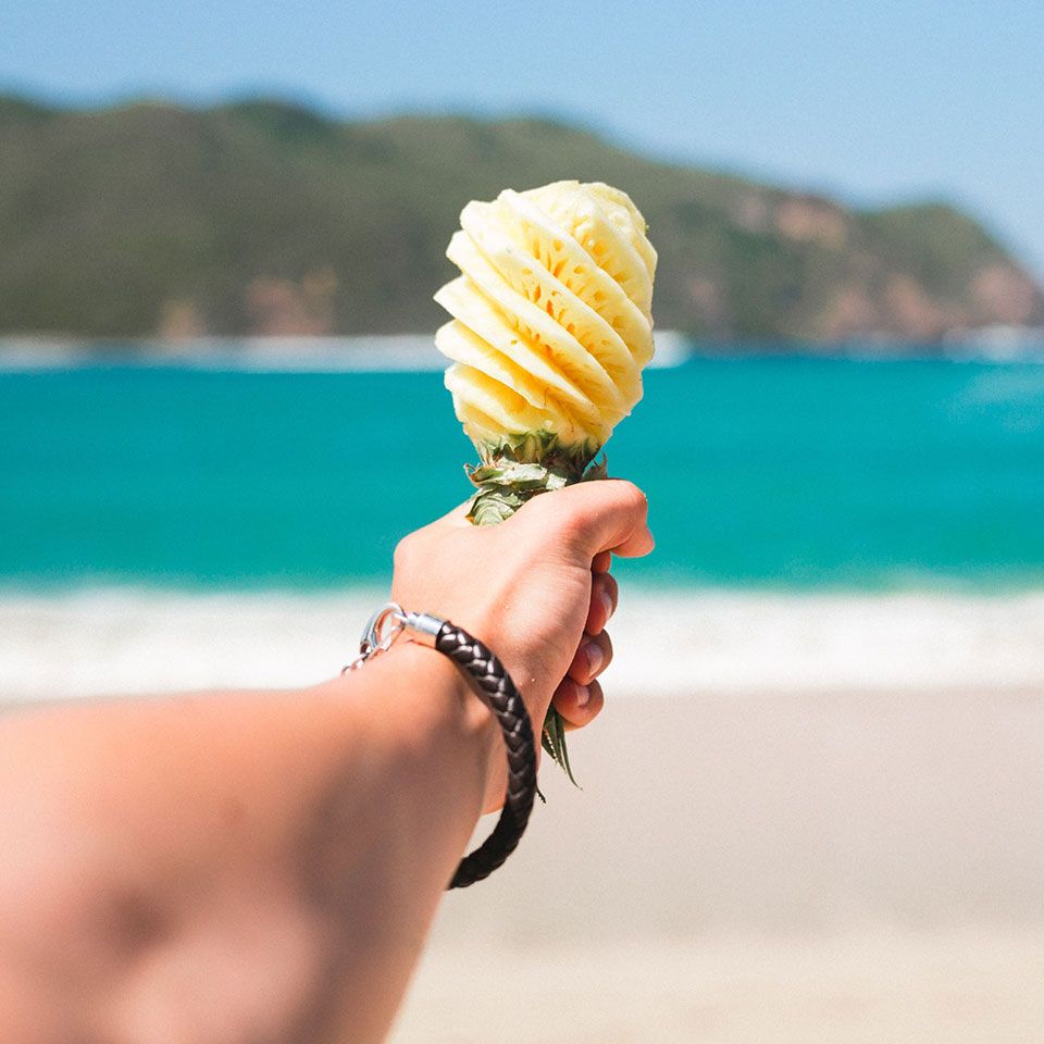 Ein ausgestreckterArm hält eine Ananas, die wie ein Mikrophon geschnitzt ist, in Richtung Strand